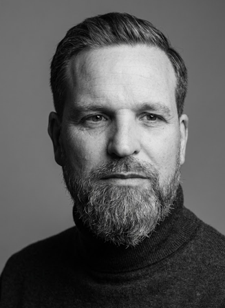 A black and white photo of a white male with moustache and a beard, wearing a dark top - looking into the camera