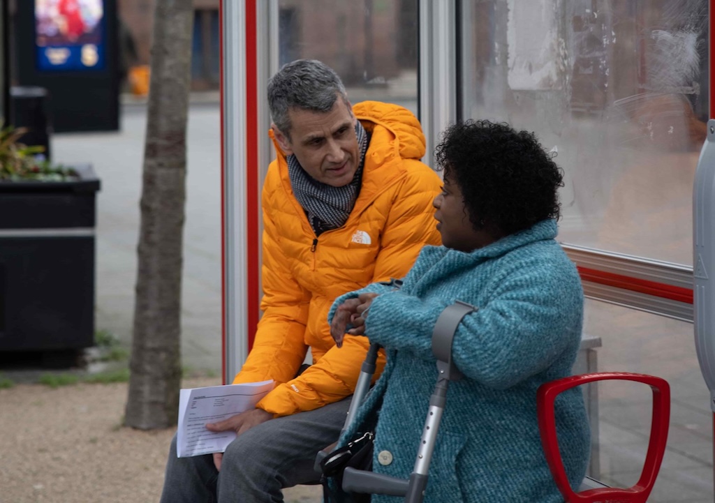 Man in orange coat talks to women in blue coat at bus shelter. A tree and flower pot are in the background of the image.